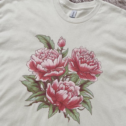 Stylish t-shirt with a drawing of Japanese tattoo art style pink peony flowers. The drawing of the pink peonies stands out against the t-shirt fabric. The drawing of the flowers is intricate and detailed, with a variety of petals and leaves. The colors of the flowers range from light pink to a deep pink. The leaves are dark green in color. 