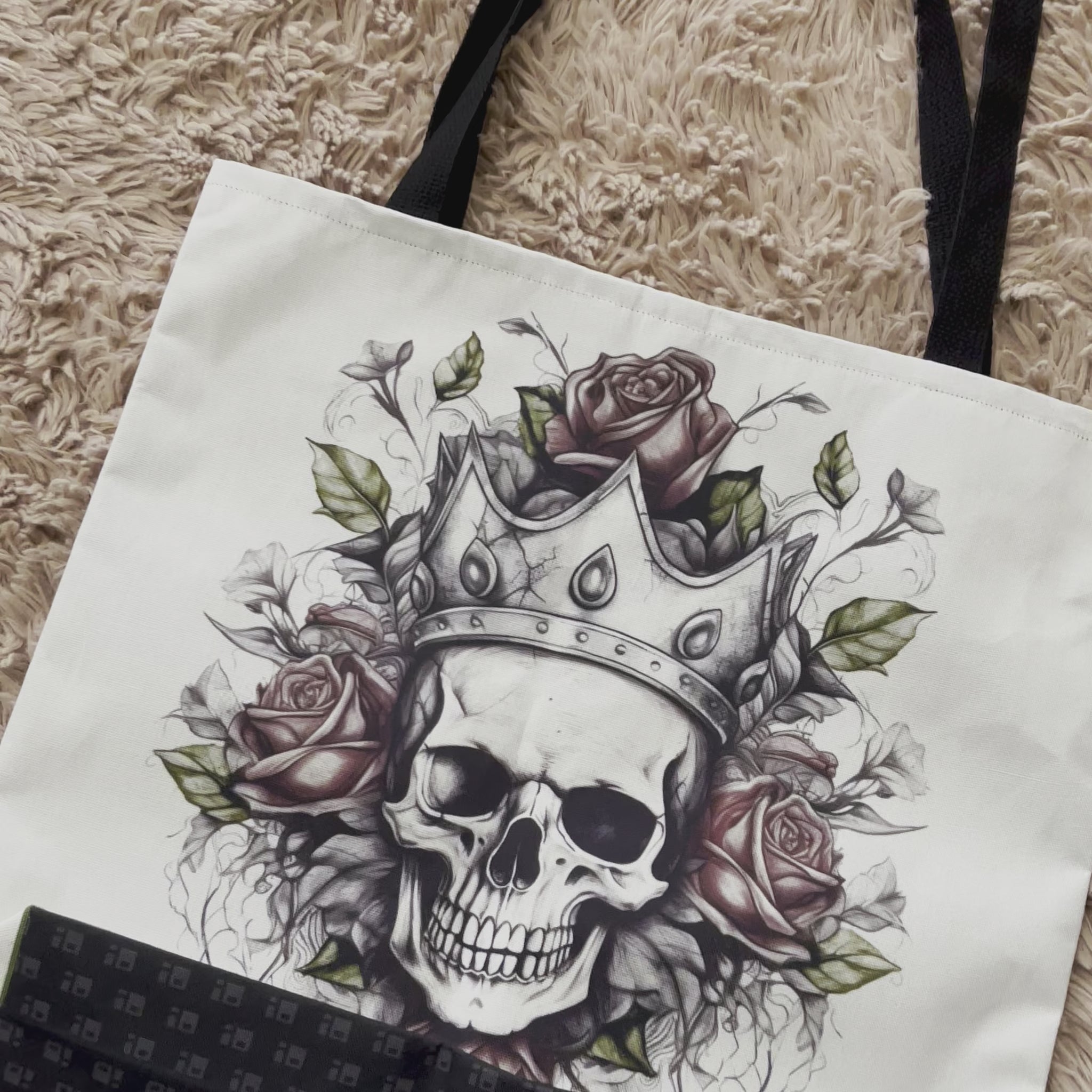 Video of a white tote bag featuring an intricate and detailed tattoo art image of a skull with a crown and red roses in muted colors. The base of the skull is surrounded by white, while the top is black and features a crown made up of intricate details. The roses are placed symmetrically on either side of the crown, as well as the top and bottom and the stems of the roses fade out into white. The bag is complete with a black strap that sits on top of the white background.